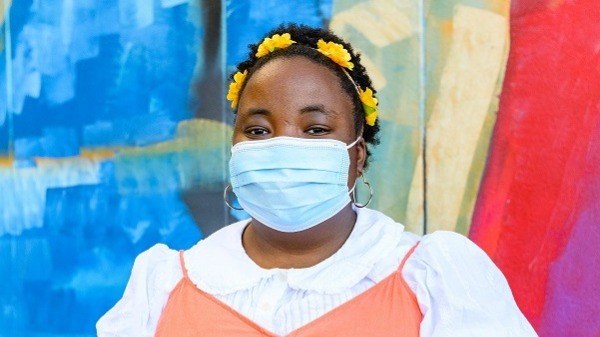 Young Black woman with yellow flower headband and short natural hair, smiling with her eyes and wearing a facemask. She stands with her arms folded in an orange dress with white collar and sleeves, outside, in front of a colorful mural.