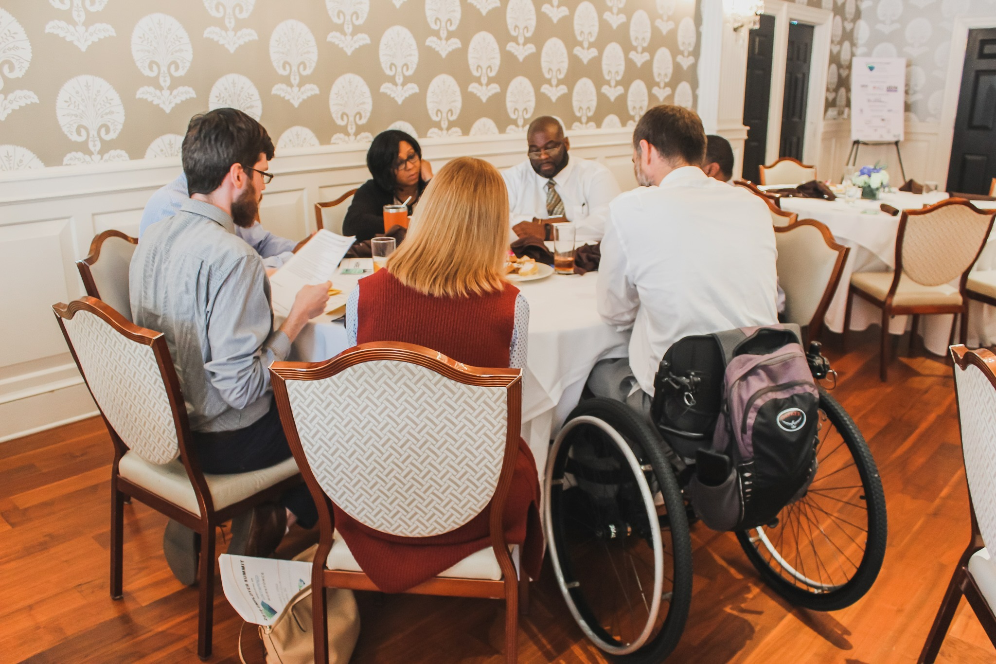 Seven people, including someone in a wheelchair, sitting around a table talking.