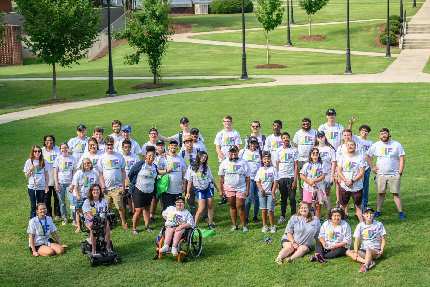 A group photo of YLF participants and staff smiling at the camera. Everyone is wearing a YLF tshirt with multi-colored YLF letters. The group is standing on a grassy area outside at Presbyterian College with small trees and lampposts in the background.