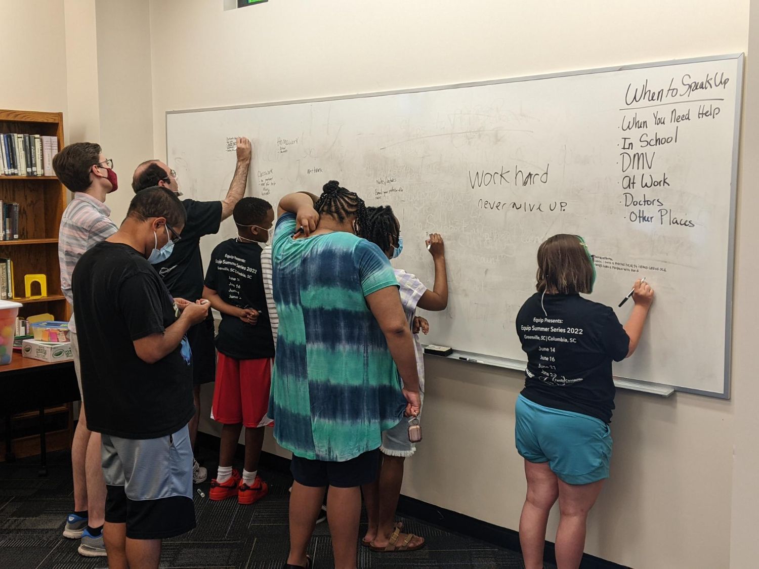 A group of young adults with disabilities who are wearing face masks stand facing a white board at the Equip Summer Series. Several of them write encouraging messages on the board such as work hard, and never give up. Others stand behind them waiting their turn to write messages on the board.