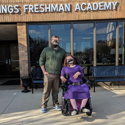 Photo of Michael, a white man with glasses and short dark hair, smiling behind a black surgical mask with an ASL window, stands next to Carrie, a white woman with shoulder length reddish brown hair, smiling behind a black surgical mask with an ASL window and a purple dress, sitting in a black power wheelchair. 'Boling Springs Freshman Academy' is written on the building behind them.