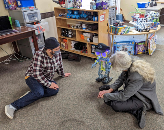 Photo of Dr. Dawson, a white man with grey hair in a ponytail, wearing a dark grey jacket and jeans, and Holden, a white man with short brown hair, wearing a plaid shirt and dark jeans, sitting on the floor beside a purple robot with a black hat on its head