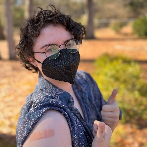 Able staffer, e.k. hoffman, poses for the camera with their booster shot band aid on their right arm while smiling to the camera behind a mask.