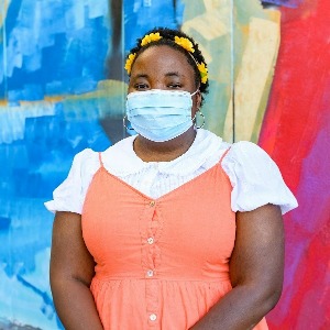 Young Black woman with yellow flower headband and short natural hair, smiling with her eyes and wearing a facemask. She stands with her arms folded in an orange dress with white collar and sleeves, outside, in front of a colorful mural.