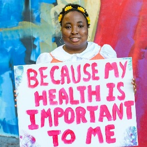A young Black woman with disabilities holding a sign that reads, ‘Because my health is important to me.’ She has yellow flowers in her hair and is wearing an orange dress while standing in front of a mural outside.
