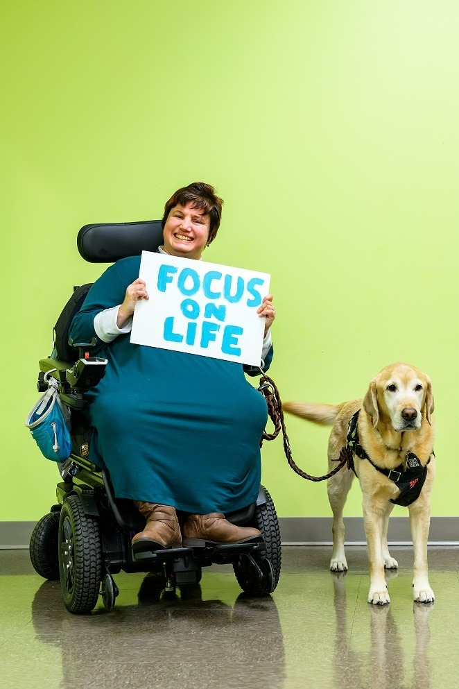 Photo of Dori Tempio, a white woman in a power wheelchair with a service dog wearing a dark blue dress, holding a hand painted sign that reads, 'Focus on life.'