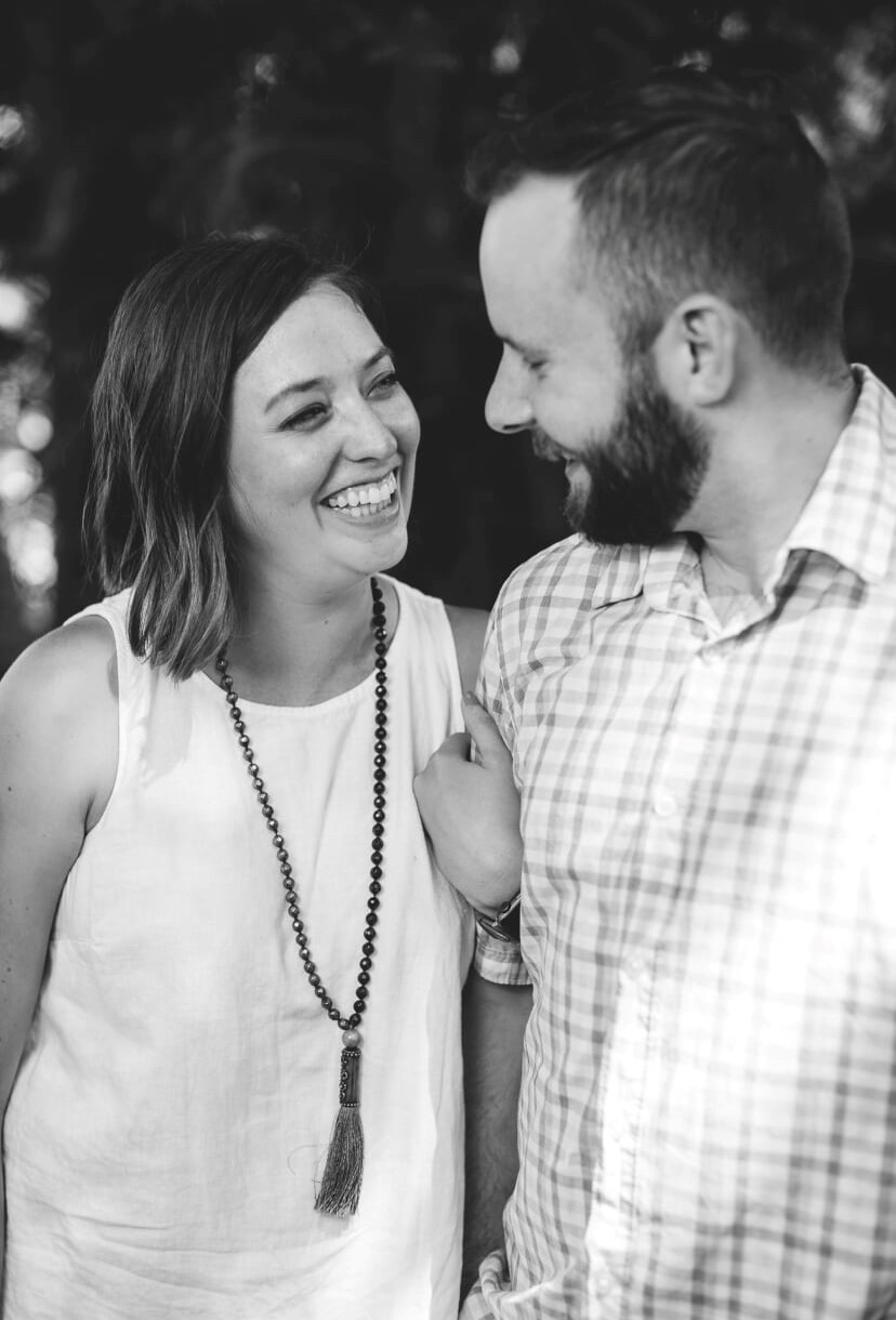 Black and white photo of Paige, a white woman with shoulder length hair, and her husband, a white man with cropped hair and a beard. Paige is holding her husbands arm on his right side and smiling wide as she looks at him. He is smiling looking toward her, with only his profile visible.