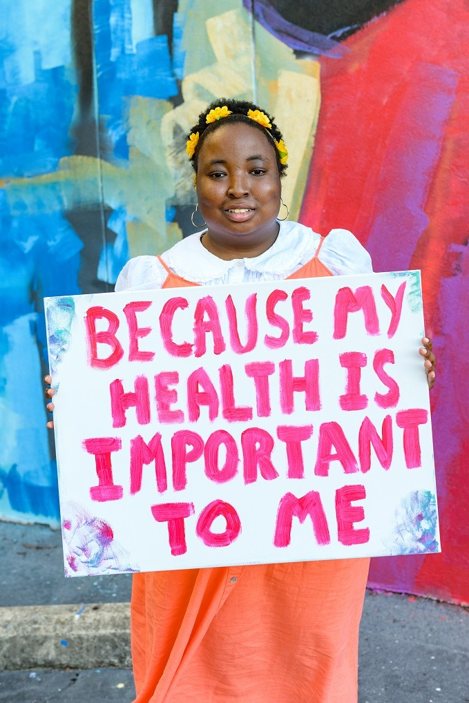 A young Black woman with disabilities holding a sign that reads, ‘Because my health is important to me.’ She has yellow flowers in her hair and is wearing an orange dress while standing in front of a mural outside.