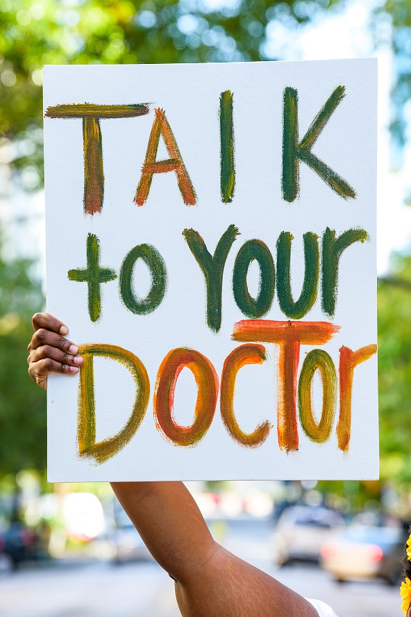 Photo of hand painted sign being held by a person with dark skin, with only their arm visible. The sign reads, 'Talk to your doctor.'