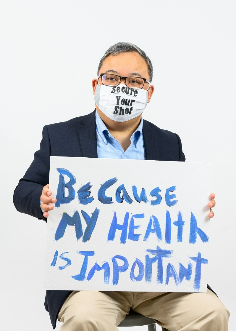 Un hombre de linaje asiático tiene un póster que dice, ‘Porque mi salud es importante’ mientras lleva una mascarilla que dice, ‘Obtener tu vacuna.’ Él tiene pelo cortado y gris y está llevando gafas y un blazer.