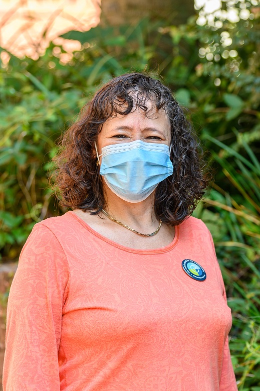 Hispanic woman with a mask on smiles. She has curly brown hair and is wearing a button that reads, 'I'm vaccinated for my community.'