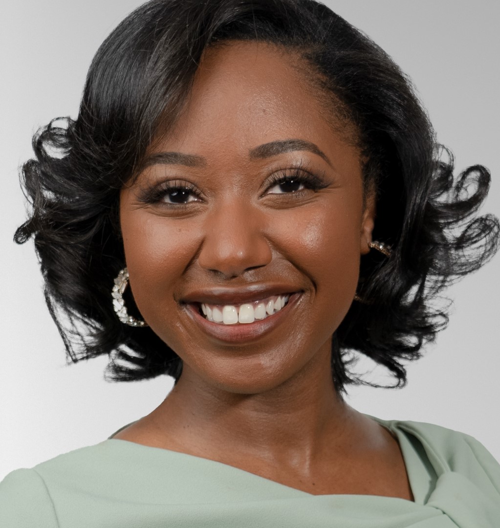 Photo of Kayland, a Black woman with short hair wearing hoops and a green dress, smiling.