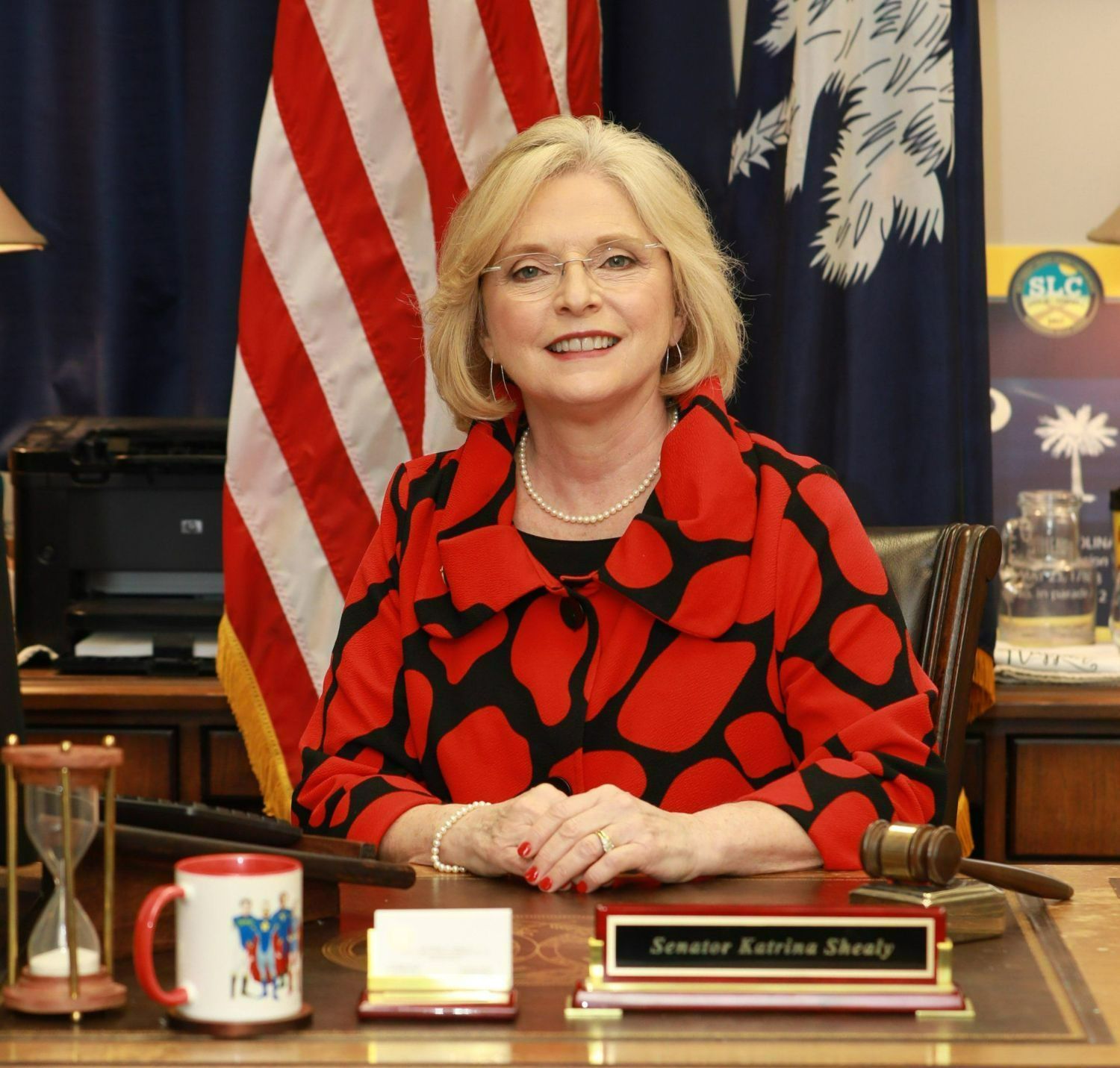 Photo of Senator Shealy, a white woman wearing a black and red blazer while sitting at a desk in front of the USA and SC flags.