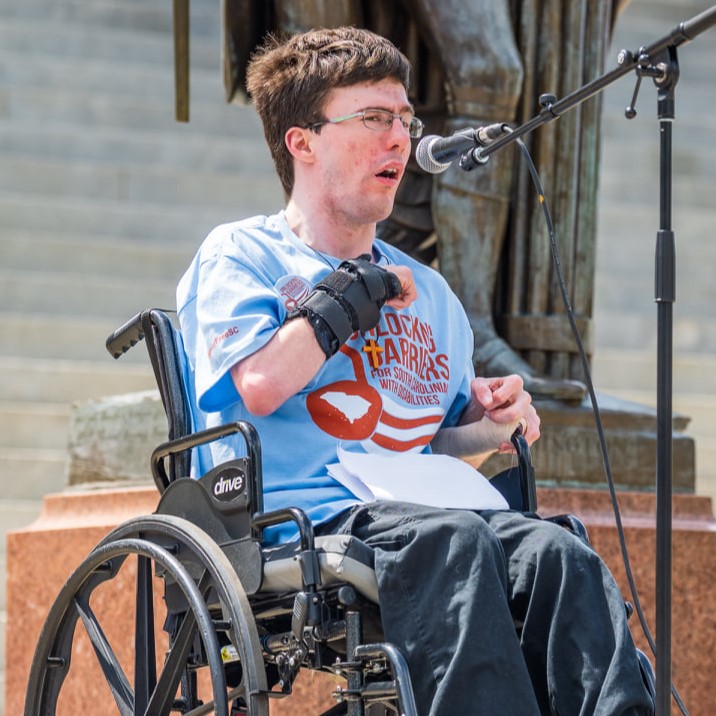 Photo of Cody, a white man using a wheelchair while speaking into a microphone at Advocacy Day.