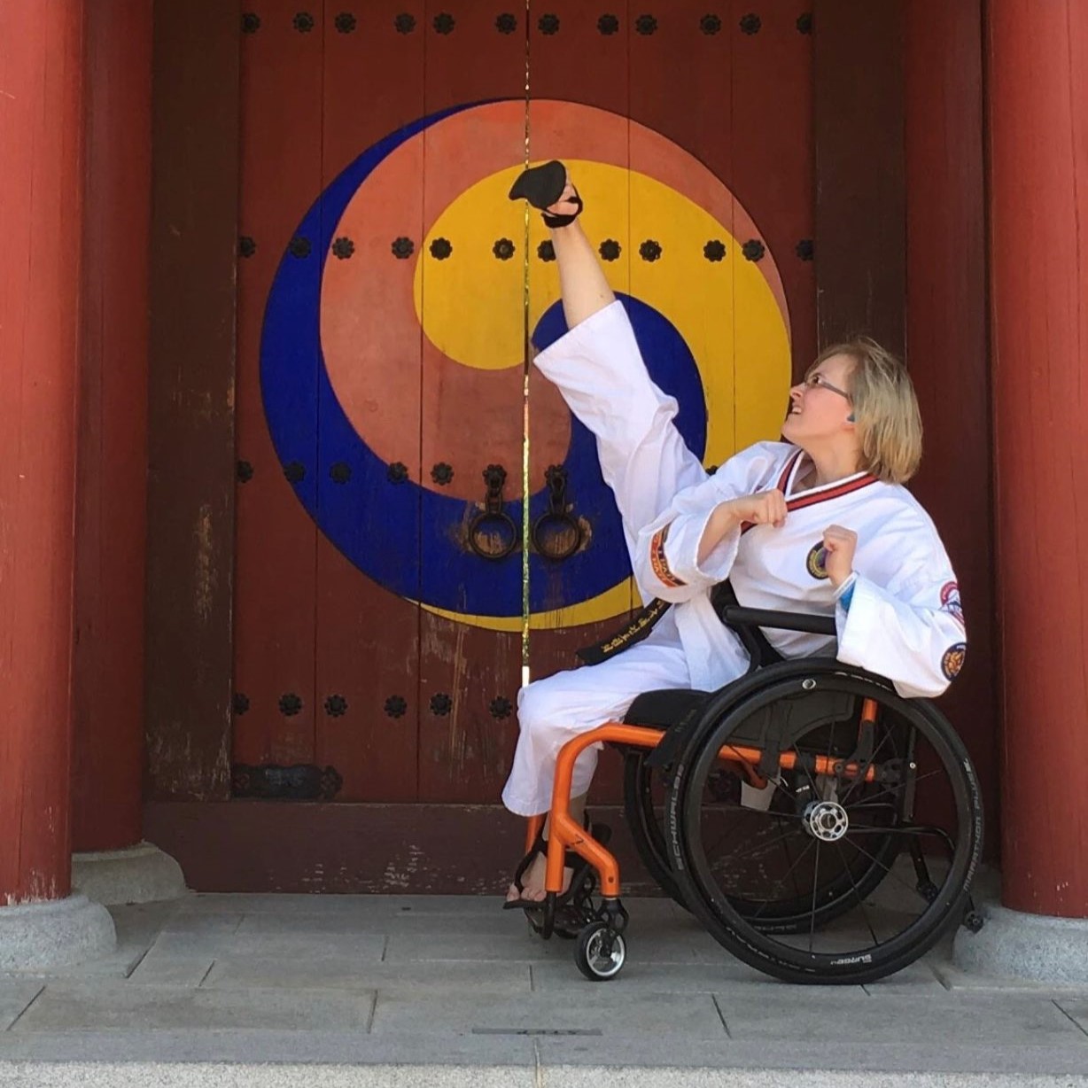 Photo of Meghan, a white woman in a wheelchair wearing a martial arts outfit and kicking her leg high into the air.