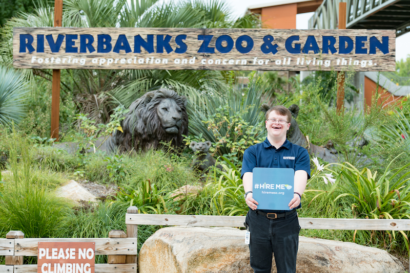 Karl Hoecke standing in front of the Riverbanks Zoo and Garden sign holding a Hire Me SC sign.