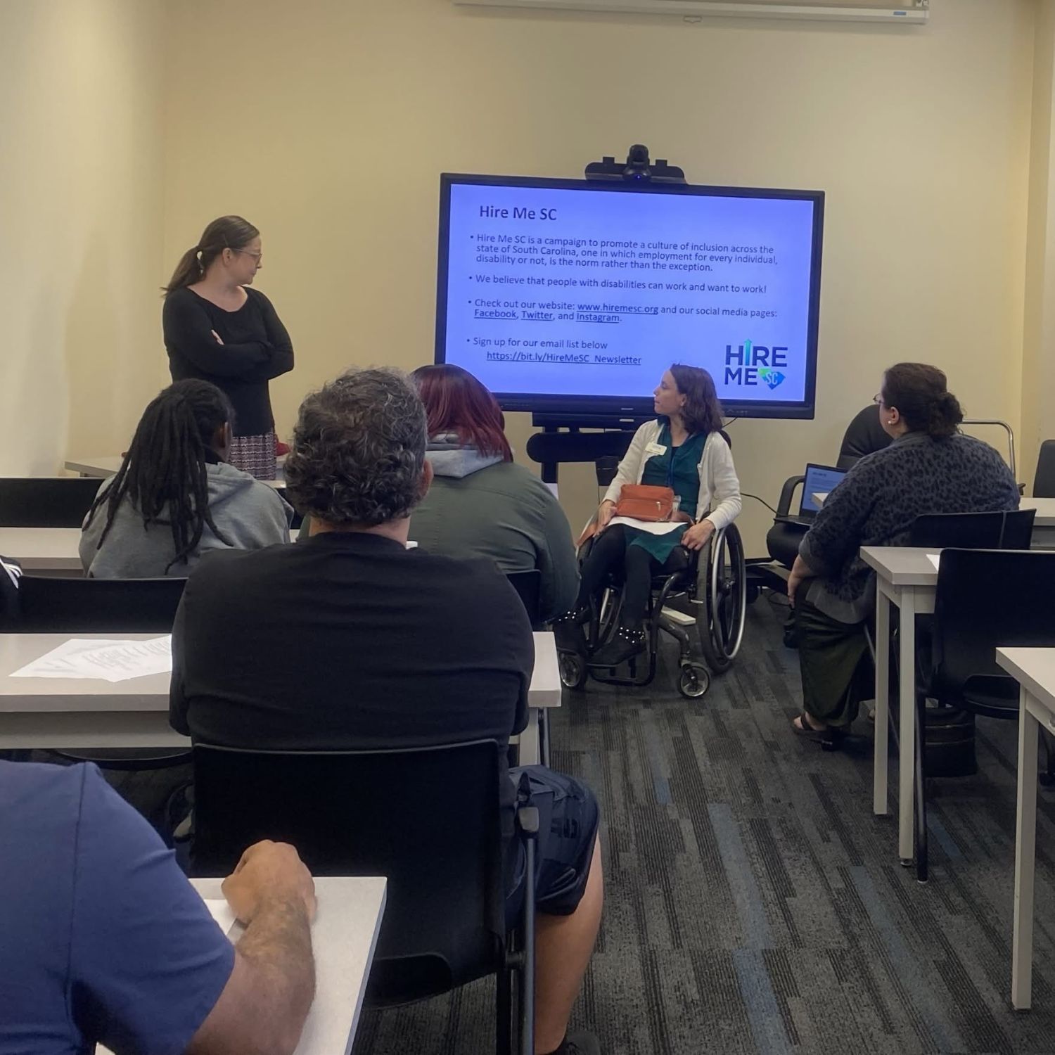 Photo of Julia, a white woman in a wheelchair, and partners presenting, with Brittany, a white woman seated in front of a laptop, operating the presentation screen. The photo is taken from the back of the room, with the backs of participants in the foreground.