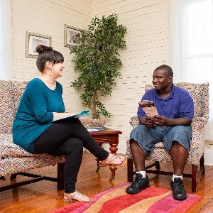 A white woman sitting with a young Black man with a developmental disability, holding materials and talking.