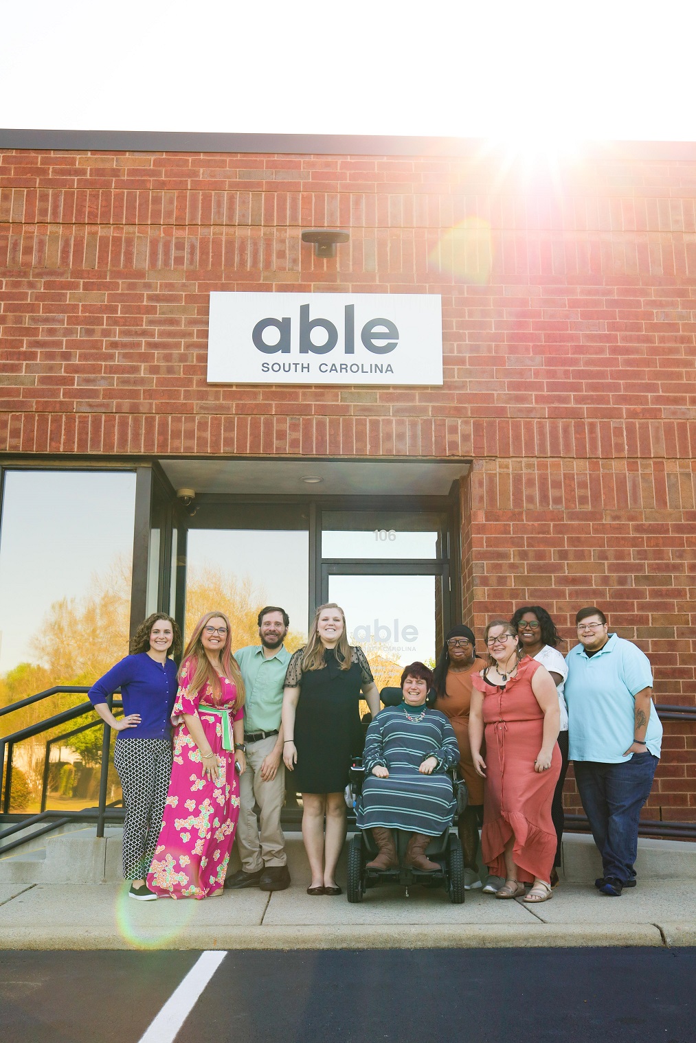 Able SC staff and consumers, diverse in age, race, gender, and disability pose outside for a photo in front of their office, with Able SC sign visible.