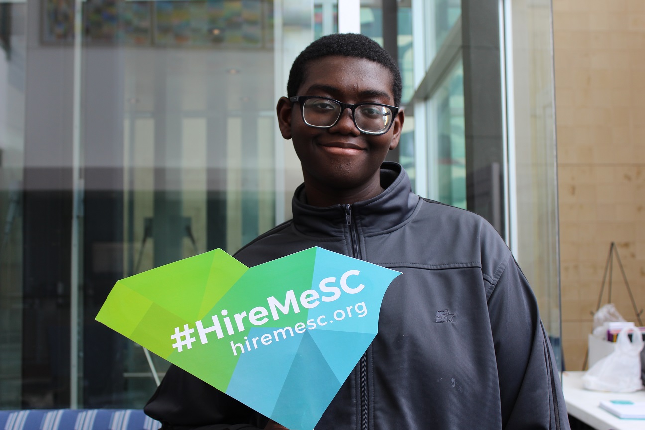 Photo of a young Black man holding a sign in the shape of South Carolina that reads 'Hire Me SC.'