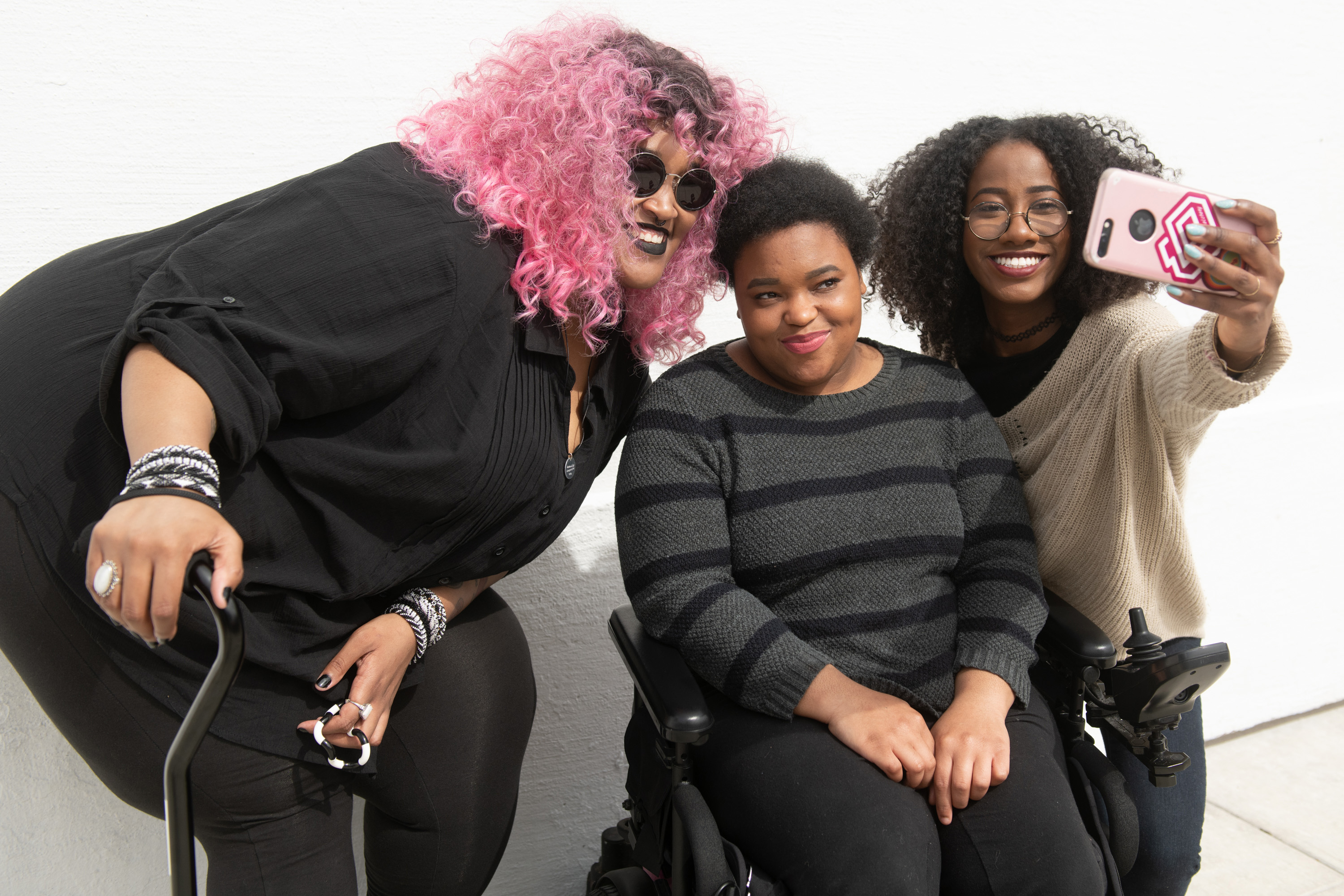 A tight crop of three Black and disabled friends (a non-binary person with a cane and tangle stim toy, a non-binary person sitting in a power wheelchair, and an invisibly disabled femme) smiling and taking a cell phone selfie together.