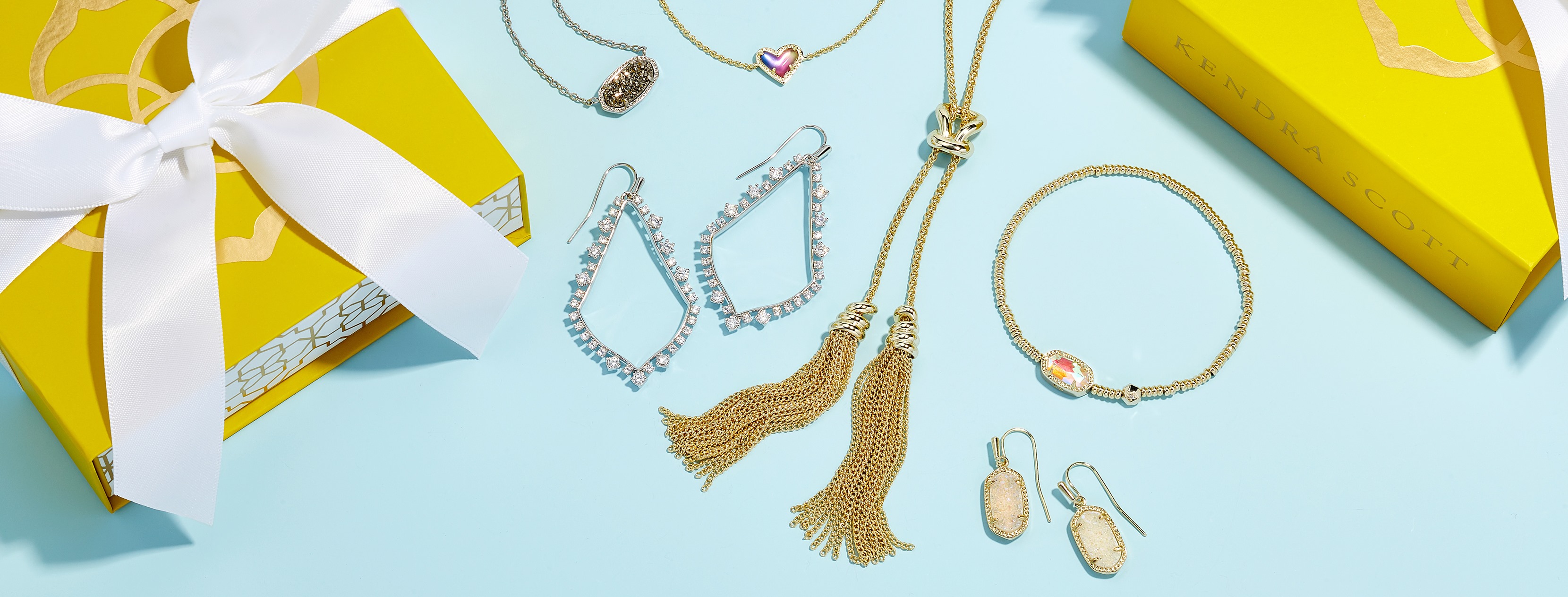 Photograph of two bright yellow gift boxes with white bows on an aqua background framing the right and left sides of the picture. In the center are several pieces of jewelry; gold chain necklace with glittery pendant, gold chain necklace with pink heart pendant, tear drop shaped gold earrings surrounded with clear crystals, gold chain necklace with doubled gold chain tassels through a bolo style loop, gold beaded bracelet with orange pendant, and small gold earrings with oval opal stones.