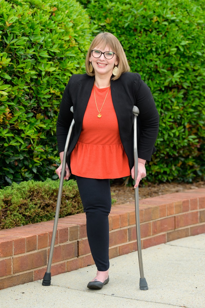 Kimberly is a white woman with shoulder length blonde hair with bangs. She is wearing brown rounded rim glasses, gold teardrop earrings, and gold necklace with a red shirt, black blazer, black pants, and gray ballet flat. She has one leg and uses crutches to assist her as she stands on a sidewalk in front of greenery, smiling for the camera.