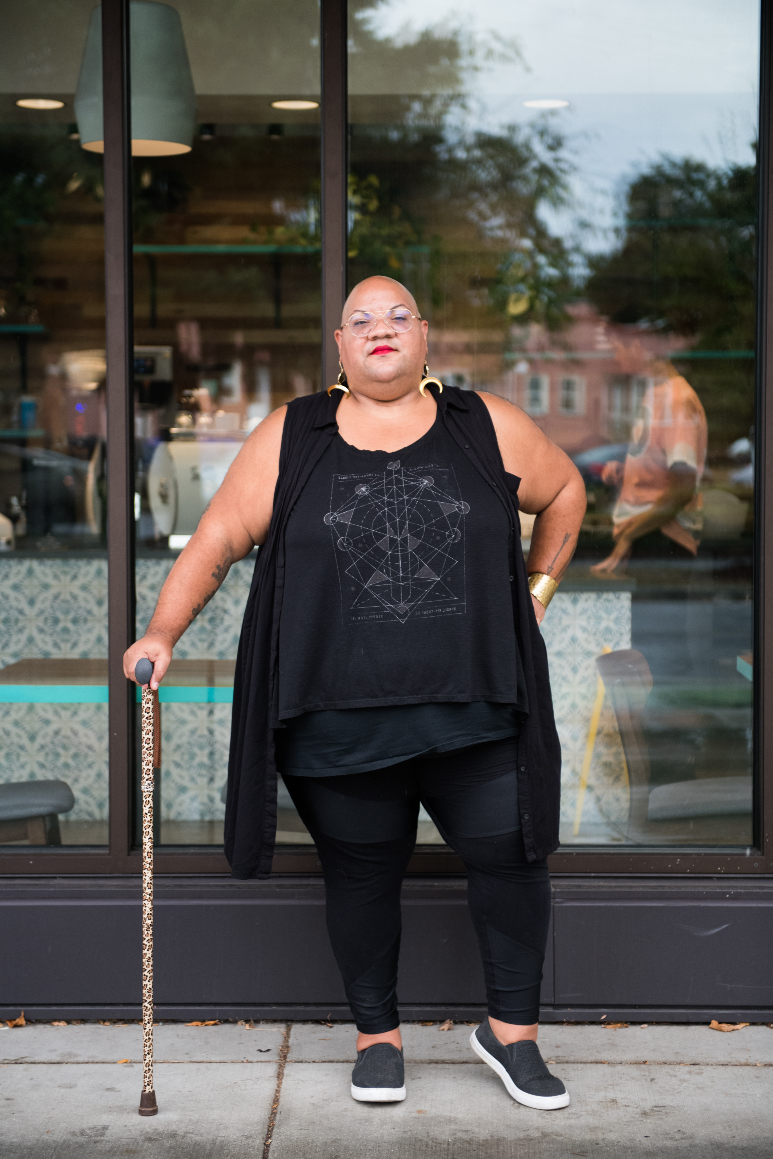 A Black non-binary person stands casually outside a cafe while leaning on their leopard print cane. They are dressed in all black and have a shaved head, glasses, and a red lip, along with moon earrings and a gold bracelet.