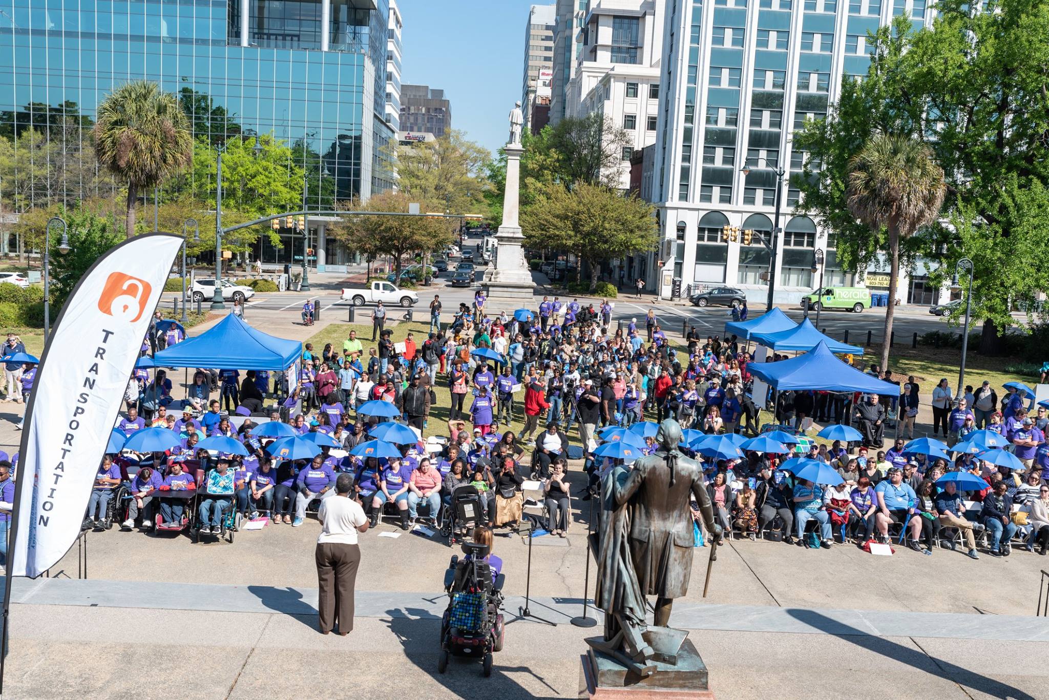 Photo of SC Statehouse grounds taken from the steps of the statehouse looking down at the crowd at Advocacy Day in 2019. A speaker is centered on the steps and people in purple shirts with blue umbrellas are watching.