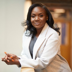 Color photo of Dr. Corett, a young Black woman with long hair wearing a white lab coat. She poses leaning on a wall with her long orange nails visible as she claps her hands together.