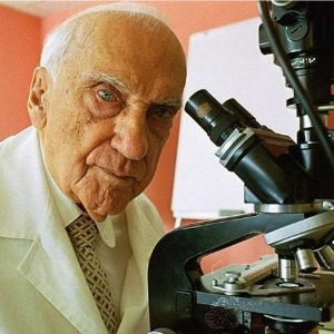 Color photo of Dr. Convit, a Latino man older in age with white hair wearing a white lab coat with a tie, posed next to a microscope.