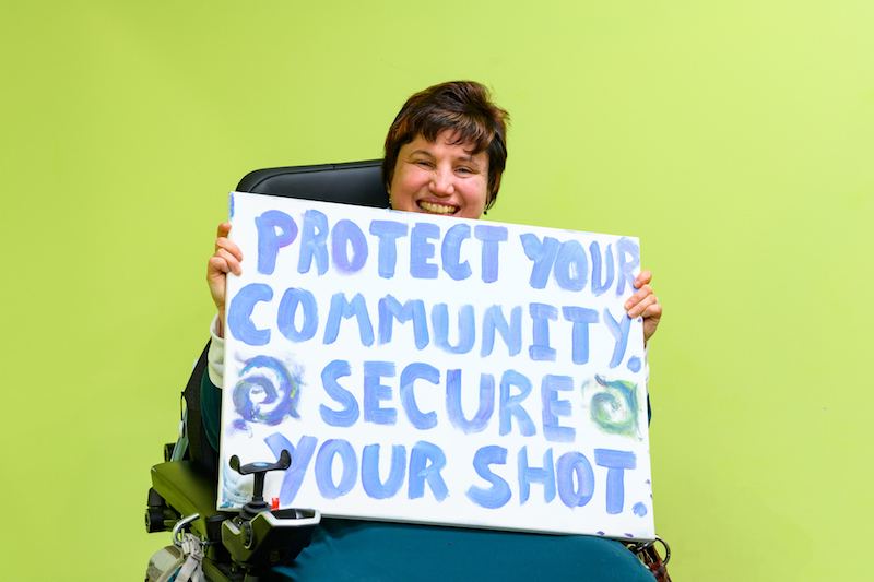 Photo of Dori, a White female using a power wheelchair. Dori has short brown hair and smiles at the camera, holding a sign that says 'Protect Your Community, Secure Your Shot'.