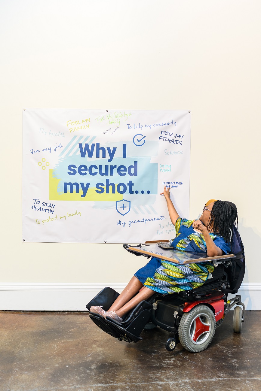 Ebony, a Black woman with braids wearing classes and using a power wheelchair with a tray, points to a banner that reads, 'Why I secured my shot...' and contains positive messages about why people with disabilities got vaccinated.