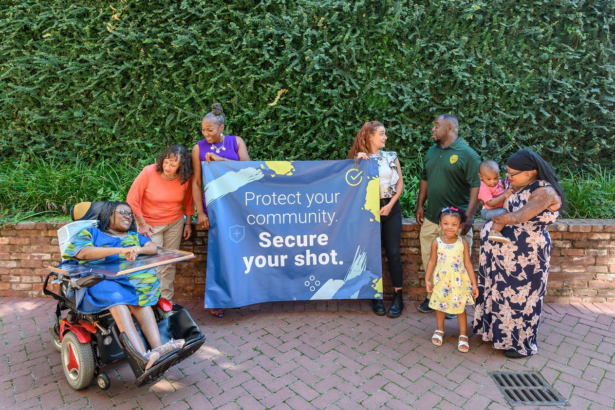 Disability Vaccine Access Leaders gathered in a group outside, holding up campagin banner reading, 'Protect your community, secure your shot.' From left to right, a young Black woman using a power wheelchair, a middle aged Hispanic woman standing and smiling, a senior Black woman with gray hair holding the banner and smiling. a young white woman with red hair holding the banner and standing while talking to her neighbor, a young adult Black man, a young Black toddler standing next to her mother, a Black woman with burn scars holding her infant Black son.