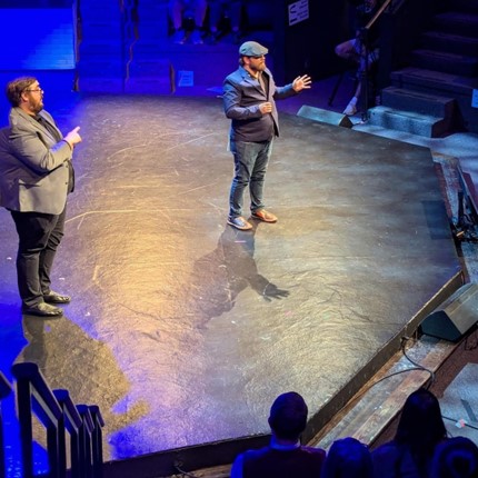 A photo of Holden, a white man with a brown beard, on the stage speaking alongside an ASL interpreter. He is surrounded by blue light and standing in a white spotlight. He is wearing a hat and suit jacket.