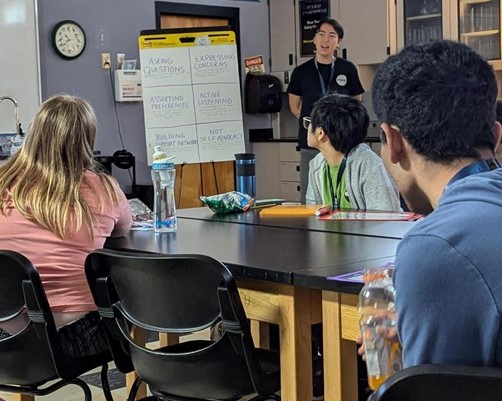 This image shows a classroom setting with a diverse group of students seated at tables, attentively facing a speaker at the front of the room. The speaker is standing near a flip chart with several topics listed, such as ‘Asking Questions,’ ‘Expressing Concerns,’ ‘Asserting Preferences,’ ‘Active Listening,’ ‘Building Support Networks,’ and ‘Not Self-Advocacy.’ The classroom has various educational materials, including a periodic table of elements, cabinets with glassware, and a clock on the wall. The atmosphere appears to be inclusive and focused on learning about communication and advocacy skills.