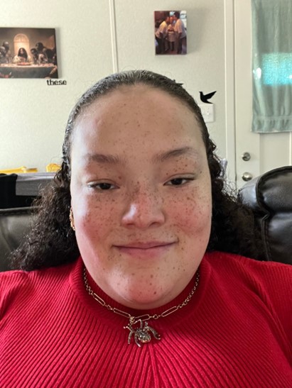 a headshot image of Dajha, a Black woman with freckles and mid length wavy brown hair. She is smiling slightly and wearing a red sweater. She is also wearing a metallic necklace with a large spider pendant